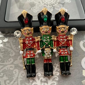 May include: A brooch featuring three toy soldier figures. Each soldier is adorned in a different color scheme: red, green, and red. They wear black hats with jewel accents and hold crystal scepters. The brooches have gold-colored details.