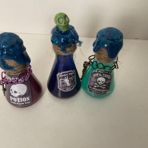 May include: Three glass potion bottles with blue, green, and purple liquid. The bottles are decorated with glitter, twine, and skull and crossbones labels. The labels read "Zombie Brew", "Unfiltered Poison", and "Potion Drink at your peril".