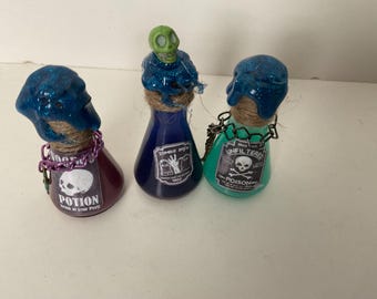 May include: Three glass potion bottles with blue, green, and purple liquid. The bottles are decorated with glitter, twine, and skull and crossbones labels. The labels read "Zombie Brew", "Unfiltered Poison", and "Potion Drink at your peril".