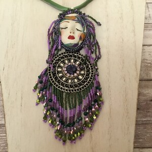 May include: A purple and green beaded necklace featuring a face pendant and a round metal charm. The face pendant has closed eyes and red lips. The round charm has a purple gemstone in the centre.