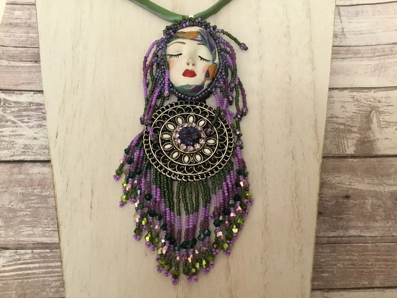 May include: A purple and green beaded necklace featuring a face pendant and a round metal charm. The face pendant has closed eyes and red lips. The round charm has a purple gemstone in the centre.