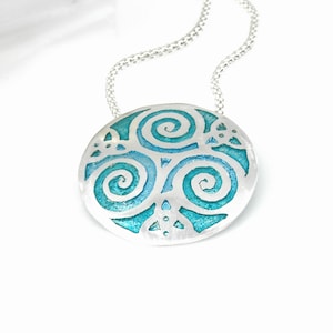 Puede incluir: Un collar de plata con un colgante redondo que presenta un diseño de esmalte azul y verde de tres espirales entrelazadas.