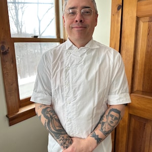 May include: A man with gray hair and glasses is wearing a white chef's coat. He has tattoos on both arms. He is standing in front of a window and a wooden door.