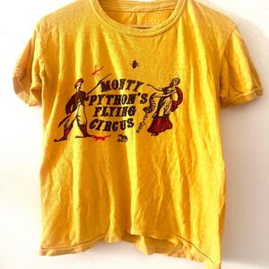 以下が含まれることがあります： 中央に「Monty Python's Flying Circus」とダークブラウンのグラフィックがプリントされた、マスタードイエローの半袖Tシャツ。赤い帽子をかぶった人物と、赤いスカートの長いドレスを着た人物のイラストが両側に描かれています。