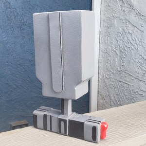 May include: A gray, geometric sculpture with a rectangular base and a boxy top. The base has black and gray stripes, and a small red accent. The sculpture is displayed on a light-colored wooden surface.