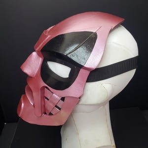 Pu&ograve; includere: Una maschera rosa e nera dal design unico, con un'area occhi nera e un cinturino. La maschera &egrave; esposta su una testa di manichino bianca su sfondo nero. La maschera ha una finitura metallica.