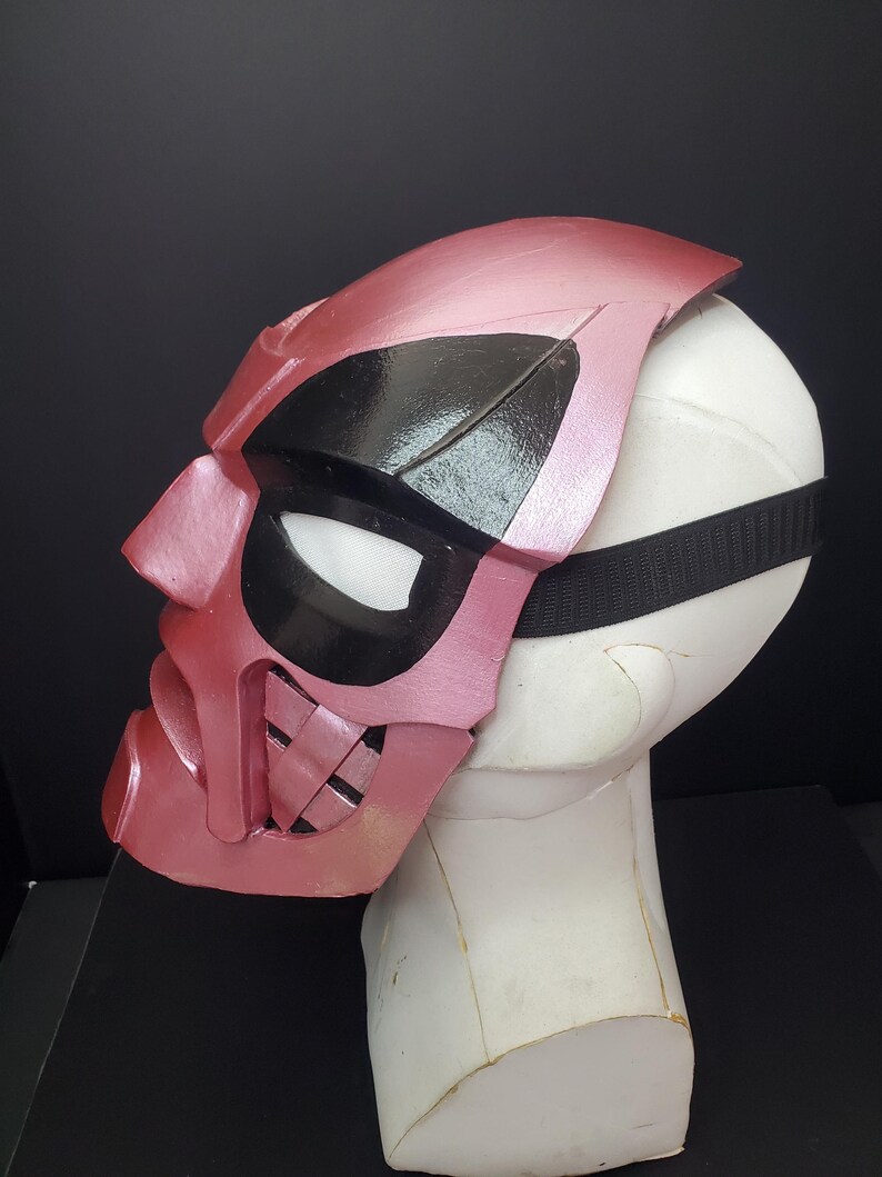 Pu&ograve; includere: Una maschera rosa e nera dal design unico, con un'area occhi nera e un cinturino. La maschera &egrave; esposta su una testa di manichino bianca su sfondo nero. La maschera ha una finitura metallica.