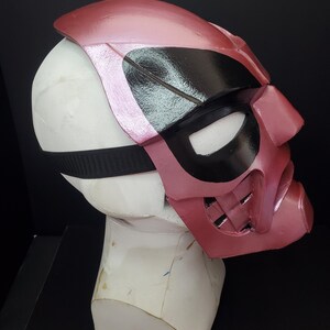 Pu&ograve; includere: Una maschera rosa e nera con un cinturino nero, esposta su una testa di manichino bianca. La maschera ha una finitura lucida e presenta aperture per gli occhi e un'area della bocca con un design strutturato. La maschera &egrave; probabilmente per cosplay o costumi.