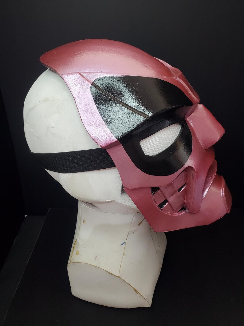 Pu&ograve; includere: Una maschera rosa e nera con un cinturino nero, esposta su una testa di manichino bianca. La maschera ha una finitura lucida e presenta aperture per gli occhi e un'area della bocca con un design strutturato. La maschera &egrave; probabilmente per cosplay o costumi.