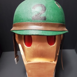 May include: A handmade, gold-colored helmet with red eye openings and a green helmet top. The green top has a black number 2 painted on it, and a brown strap wraps around the base. The helmet has a vintage, retro aesthetic.