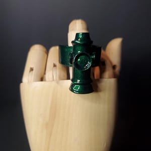 May include: A dark green ring with a metallic, glittery finish featuring a central circular emblem and a vertical, pillar-like design element. The ring has a smooth, polished surface and a wide band that wraps around the finger area.