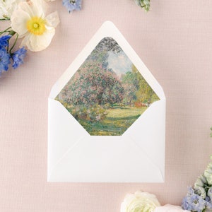Pode incluir: Um envelope branco com uma pintura de paisagem visível no interior. A pintura retrata uma cena de jardim com folhagem rosa e verde. Flores brancas e azuis são dispostas ao redor do envelope em uma superfície rosa.