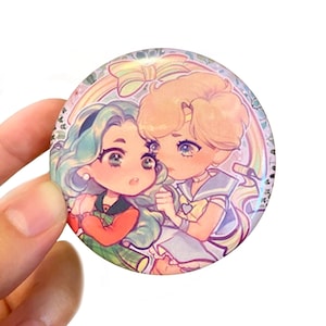 May include: A round button with a colorful illustration of two cartoon characters. The characters are a girl with green hair and a girl with blonde hair. The girls are wearing colorful outfits and are smiling. The button has a rainbow background and the text "SUSANBIN.COM" is printed at the top.