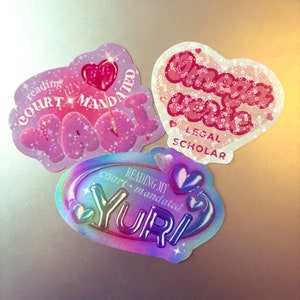 Può includere: Tre adesivi con testo e disegni a forma di cuore. Uno recita "reading my court mandated Naomi" in rosa. Un altro recita "Omega Love Legal Scholar" in rosa. Il terzo recita "reading my court + mandated Yuri" in colori iridescenti.
