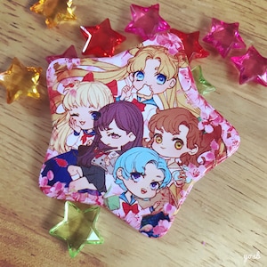 May include: A star-shaped pin with a pink and white background featuring five anime characters. The characters are a blonde girl with blue eyes, a blonde girl with pink hair, a girl with purple hair, a girl with brown hair, and a girl with blue hair. The characters are all smiling and have large eyes.