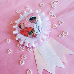 Op de afbeelding: Een roze en witte rozetbadge met een hartvormige afbeelding van twee mensen. De badge heeft een witte gefranjerde rand en roze linten. Kleine, parelachtige hartvormige versieringen zijn verspreid op een roze achtergrond.
