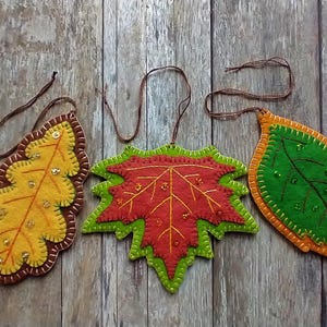 May include: Three felt leaf ornaments with embroidered details and sequins. The leaves are in shades of yellow, orange, red, and green.