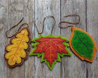 以下が含まれることがあります： 刺繍とスパンコールで飾られたフェルトの葉のオーナメント3つ。葉は黄色、オレンジ、赤、緑の色合いです。