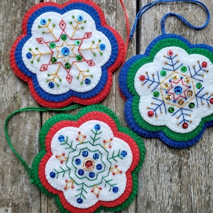 Puede incluir: Tres adornos de copo de nieve de fieltro con ribetes azules, verdes y rojos. Cada copo de nieve está decorado con hilo dorado y plateado, lentejuelas y cuentas.