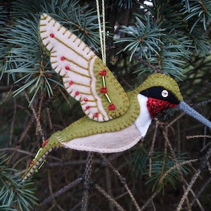 Puede incluir: Un adorno de colibrí de fieltro con detalles en verde, beige y rojo. El colibrí está en vuelo con las alas extendidas. El adorno está decorado con pequeñas cuentas rojas y verdes.