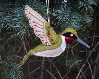Puede incluir: Un adorno de colibrí de fieltro con detalles en verde, beige y rojo. El colibrí está en vuelo con las alas extendidas. El adorno está decorado con pequeñas cuentas rojas y verdes.