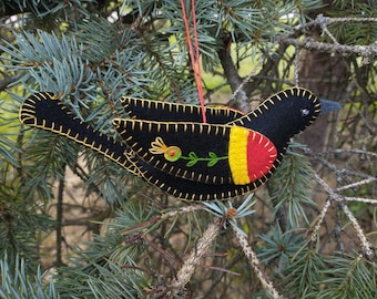 Puede incluir: Un adorno de pájaro de fieltro negro con detalles bordados en amarillo, rojo y verde. El pájaro está colgado de una rama de pino.