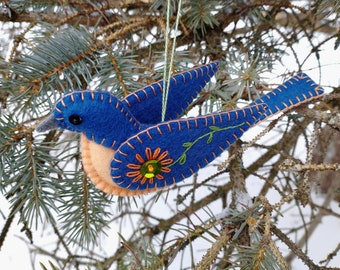 Puede incluir: Un adorno de pájaro de fieltro azul con bordados naranja y verde y un centro amarillo brillante. El pájaro está colgado de una rama de pino.