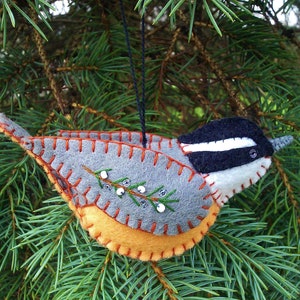 May include: A felt ornament shaped like a bird with a gray body, black head, white stripe, and orange accents. The bird has a small branch with white beads on it.
