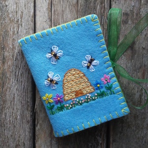 May include: A blue felt notebook with a beehive, bees, and flowers embroidered on the front. The notebook has a green ribbon attached to it.