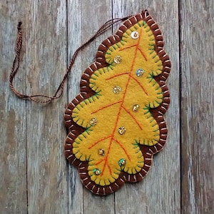 May include: A handmade felt leaf ornament in shades of yellow, brown, and green. The leaf features intricate stitching, decorative sequins, and a brown string for hanging. The ornament is designed to resemble an autumn leaf.