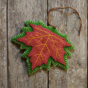 May include: A handmade felt leaf ornament in shades of red and green, with yellow stitching and embellishments. The leaf has a brown string for hanging. The ornament is set against a rustic wooden background, evoking a fall theme.