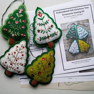 May include: Four embroidered felt Christmas tree ornaments in green, white, and yellow with brown trunks. The trees have decorative stitching and bead accents. A sewing kit with instructions is also visible.