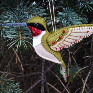 May include: A handmade felt hummingbird ornament. The bird features olive green, white, and red accents, with a long gray beak and detailed wings. The ornament is suspended by a gold string, perfect for holiday decor.