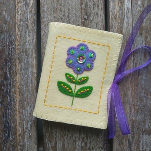 May include: A small, cream-colored felt pouch with a purple flower embroidered on the front. The flower has green leaves and yellow accents. The pouch has a purple ribbon attached to it.