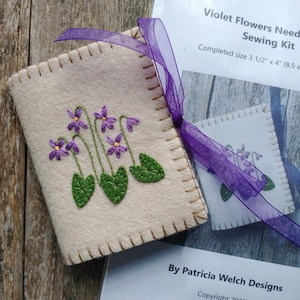 May include: A beige needlebook with embroidered purple violets and green leaves, tied with a purple ribbon. The text "Violet Flowers Needlebook Sewing Kit" is visible, with a completed size of 3.5" x 4".