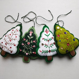 May include: Four handmade Christmas tree decorations. Each tree is a different colour: white, dark green, and light green. The trees are decorated with red beads, sequins, and embroidered details. They have brown trunks and green trim.