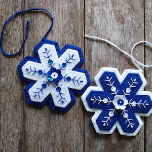May include: Two white felt snowflake decorations with blue felt trim and blue and white sequins. The snowflakes are hanging from white and blue strings.