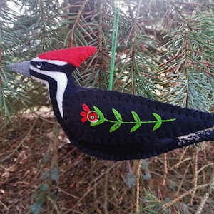 May include: A handmade felt bird ornament, resembling a woodpecker, with a red crest, black body, and white markings. The bird features green embroidered leaves and a red flower. A green string is attached for hanging.