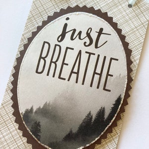May include: A brown oval-shaped tag with a white background and a black and white image of a foggy forest. The text "just breathe" is printed in black on the tag.