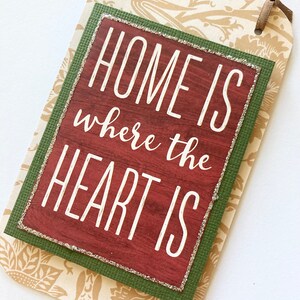 May include: A decorative tag with the text "HOME IS where the HEART IS" in white lettering. The tag has a brown ribbon and a floral pattern background. The text is on a red wooden background with a green border.