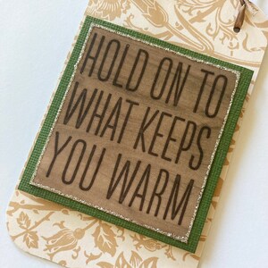 May include: A brown wooden sign with a green border and glitter trim. The sign reads "Hold on to what keeps you warm".