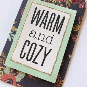 May include: A light green tag with a gold glitter border and the text "WARM and COZY" in black lettering. The tag is on a dark background with a floral pattern.