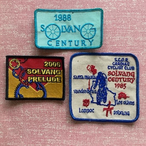 May include: Three embroidered patches featuring the Solvang Century bike race. The top patch is light blue with white lettering and shows the year 1988. The middle patch is yellow with red lettering and shows the year 2006. The bottom patch is white with blue lettering and shows the years 1985 and 2006.