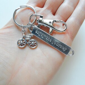 Pu&ograve; includere: Portachiavi color argento con un ciondolo a forma di bicicletta e un'etichetta rettangolare incisa con "enjoy the journey". Il portachiavi include un moschettone e un anello diviso per attaccare le chiavi. Il ciondolo della bicicletta ha raggi dettagliati.