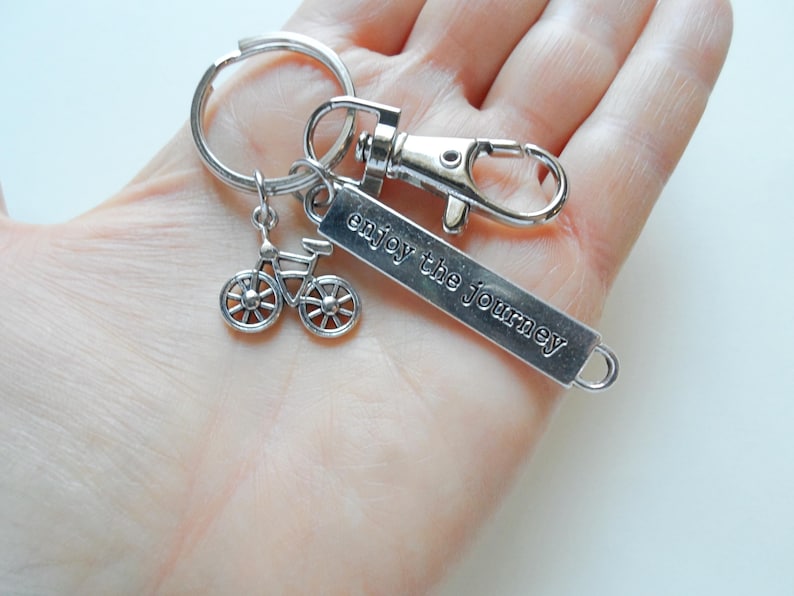 Pu&ograve; includere: Portachiavi color argento con un ciondolo a forma di bicicletta e un'etichetta rettangolare incisa con "enjoy the journey". Il portachiavi include un moschettone e un anello diviso per attaccare le chiavi. Il ciondolo della bicicletta ha raggi dettagliati.