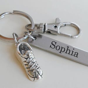 May include: Silver-colored keychain with a running shoe charm and a rectangular tag engraved with the name "Sophia". The keychain includes a split ring and a lobster clasp for easy attachment.