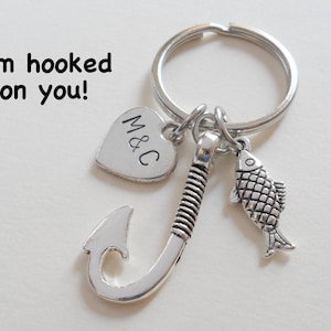 May include: Silver keychain with a fish charm, a hook charm, and a heart charm with the initials "M&C" engraved on it. The keychain has the text "I'm hooked on you!" on the background.