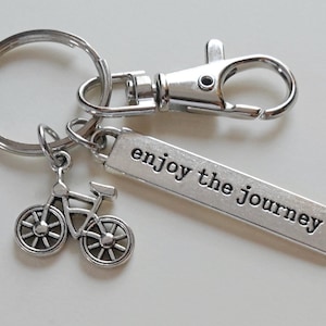 Pu&ograve; includere: Portachiavi argentato con un ciondolo a forma di bicicletta e un'etichetta rettangolare con la scritta "enjoy the journey". Il portachiavi include un anello portachiavi, un moschettone e un piccolo ciondolo a forma di bicicletta. Il testo &egrave; in un font sans-serif.