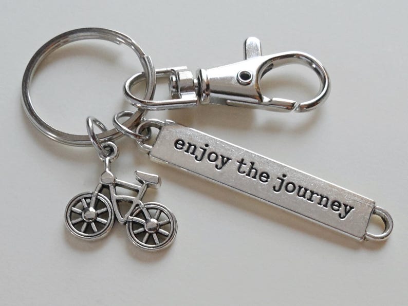 Pu&ograve; includere: Portachiavi argentato con un ciondolo a forma di bicicletta e un'etichetta rettangolare con la scritta "enjoy the journey". Il portachiavi include un anello portachiavi, un moschettone e un piccolo ciondolo a forma di bicicletta. Il testo &egrave; in un font sans-serif.
