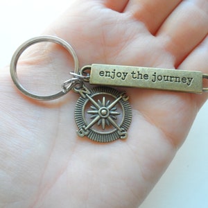 Pode incluir: Um chaveiro de b&uacute;ssola de bronze com as palavras "enjoy the journey" em uma etiqueta retangular. O chaveiro est&aacute; preso a um anel de chave prateado.
